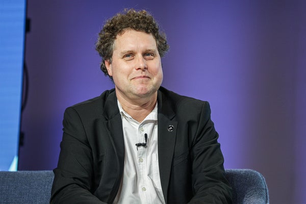 Peter Beck sitting in a blue chair on stage at a summit with a purple background